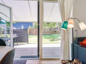 Un salon moderne doté d’une porte-fenêtre coulissante donnant sur une terrasse. On y trouve un canapé avec un coussin, une table avec des chaises et une lampe.