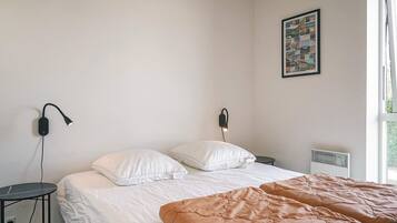 Un dormitorio con cama, mesitas de noche, lámpara de pared, radiador y ventana.