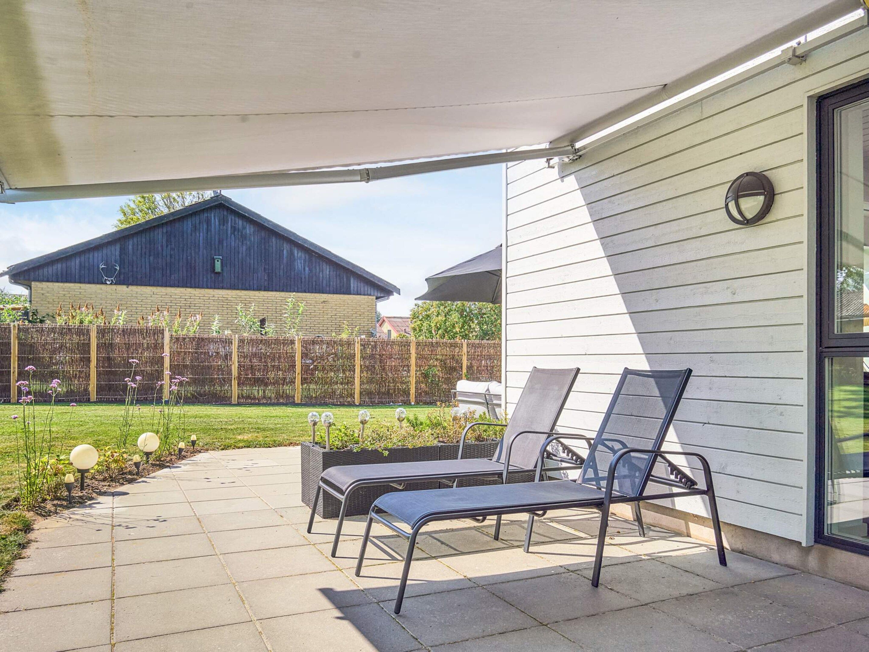 A covered patio with two lounge chairs and a table, a wooden fence, and a building in the background.