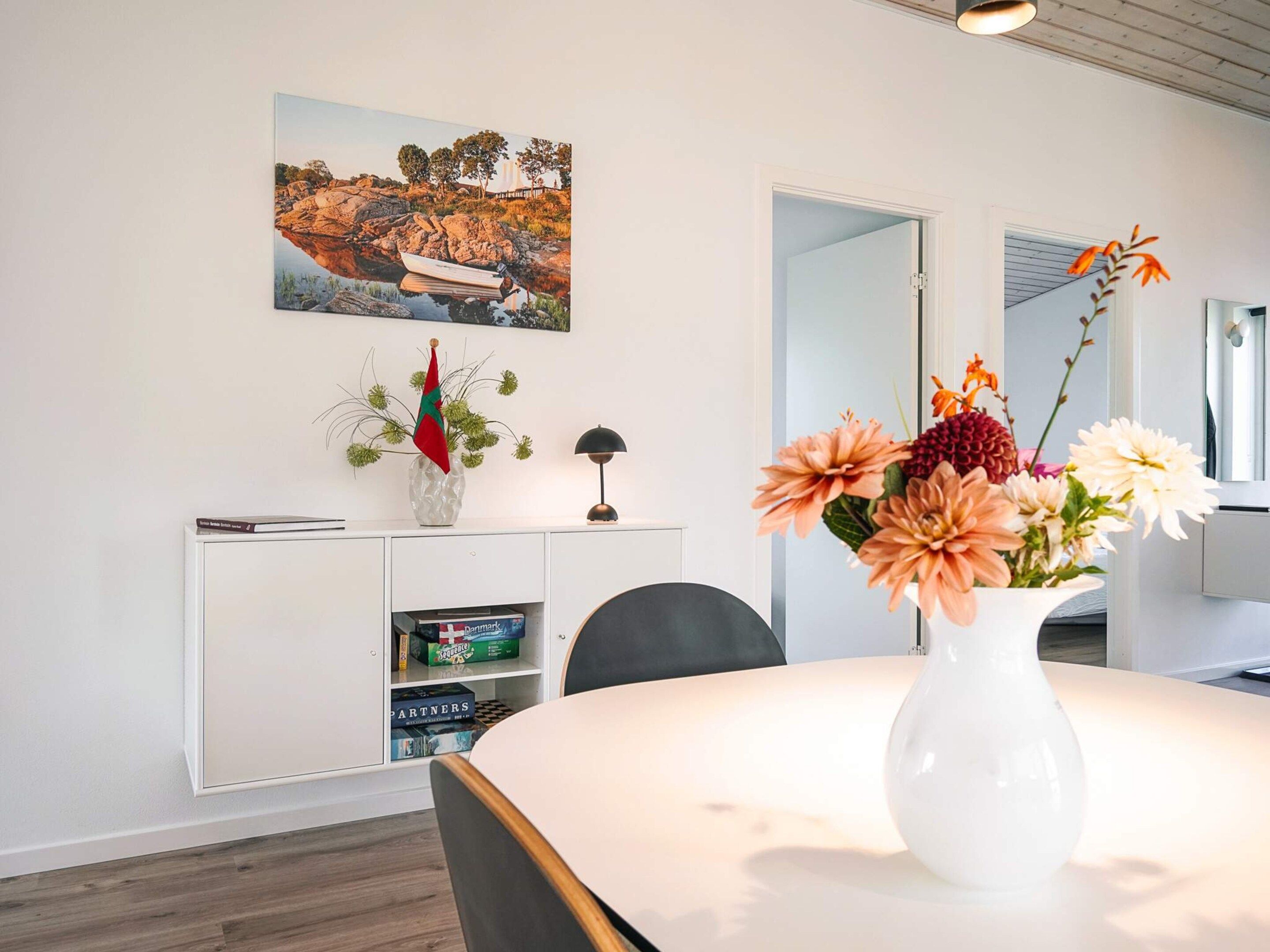 Una camera moderna con un tavolo rotondo, un mobile bianco pieno di libri, un quadro e un vaso con fiori.