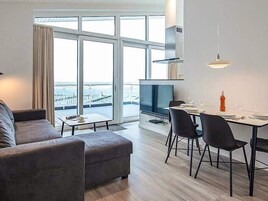 A modern living room with a sofa, dining table, chairs, a TV, and large windows offering a view of a bridge.