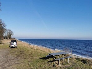 A beach with a car parked on the side of a path, a picnic table, and the ocean in the background.