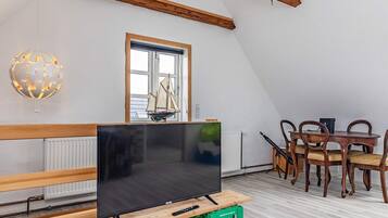 Una habitación con televisión sobre un soporte de madera, una mesa de comedor con sillas, una ventana con un modelo de velero y una lámpara con forma de globo.