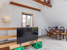 Una habitación con televisión sobre un soporte de madera, una mesa de comedor con sillas, una ventana con un modelo de velero y una lámpara con forma de globo.