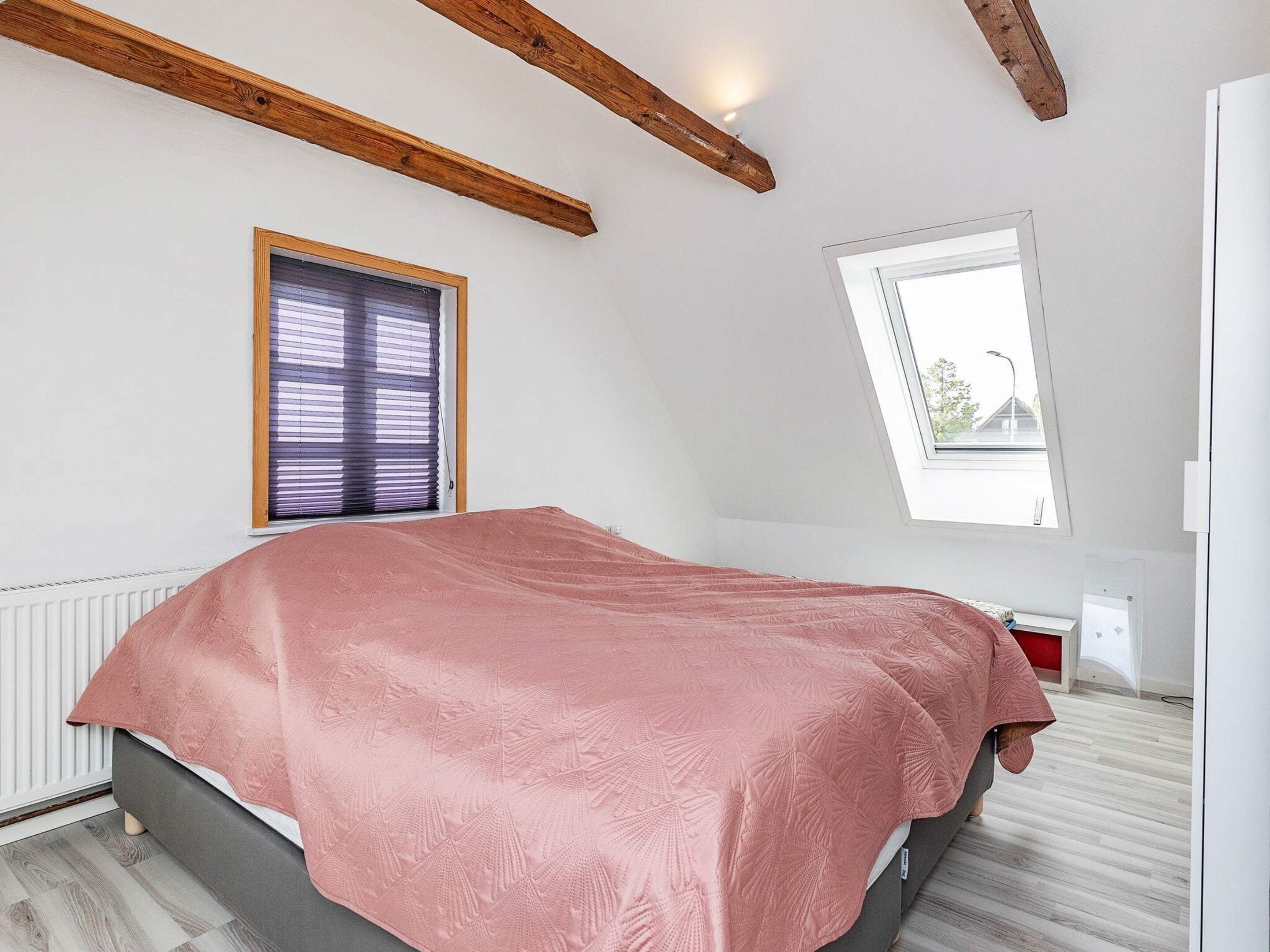 A bedroom with a bed, two windows, wooden beams, and a radiator.