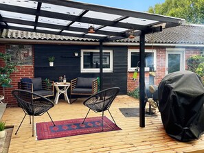 A covered outdoor seating area with a wooden deck, two black chairs, a small table, and a red patterned rug.