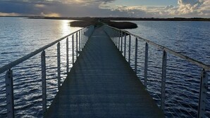 En lang bro der strækker sig ud i roligt vand med gelænder, førende til en fjerntliggende kystlinje under en delvist overskyet himmel.