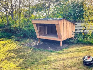 A wooden doghouse with a sloped roof, situated in a garden with trees and grass.