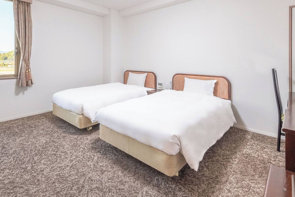 A hotel room with two single beds, a wooden headboard, a small bedside table, a desk, and a window with curtains.