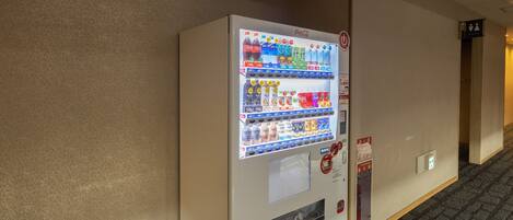 Máquina expendedora con diversas opciones de bebidas, incluyendo Coca-Cola y otras, ubicada en un pasillo con piso alfombrado y pared lisa.