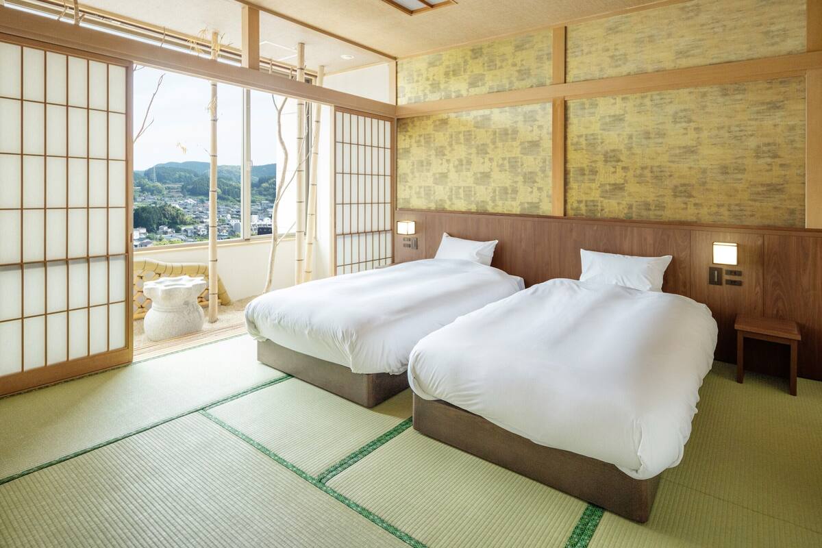 A traditional Japanese room with tatami flooring, two beds with white bedding, a view of the city, and a wooden headboard.