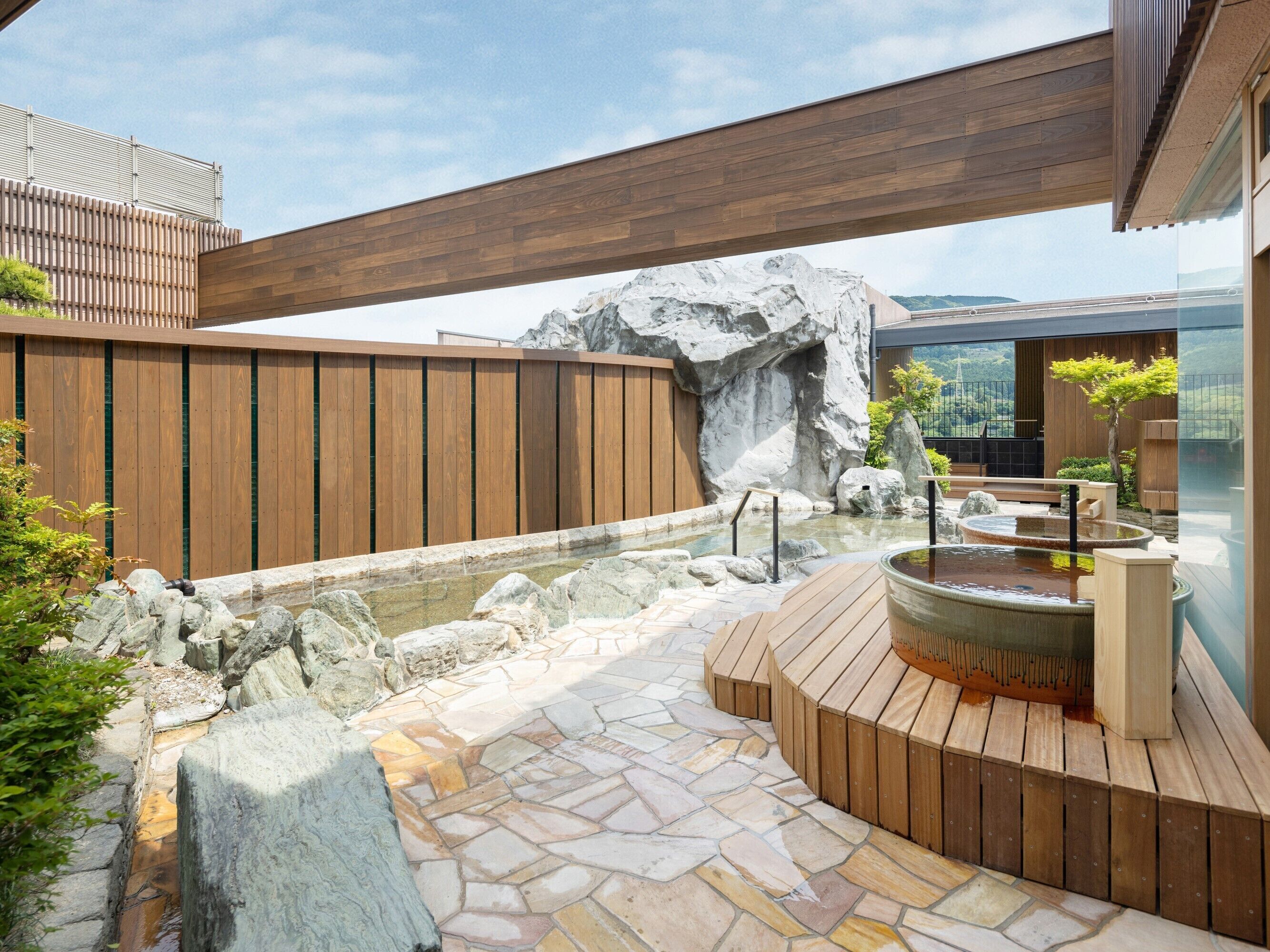 A modern outdoor spa area with a hot tub, stone pathway, wooden deck, and a large rock formation.