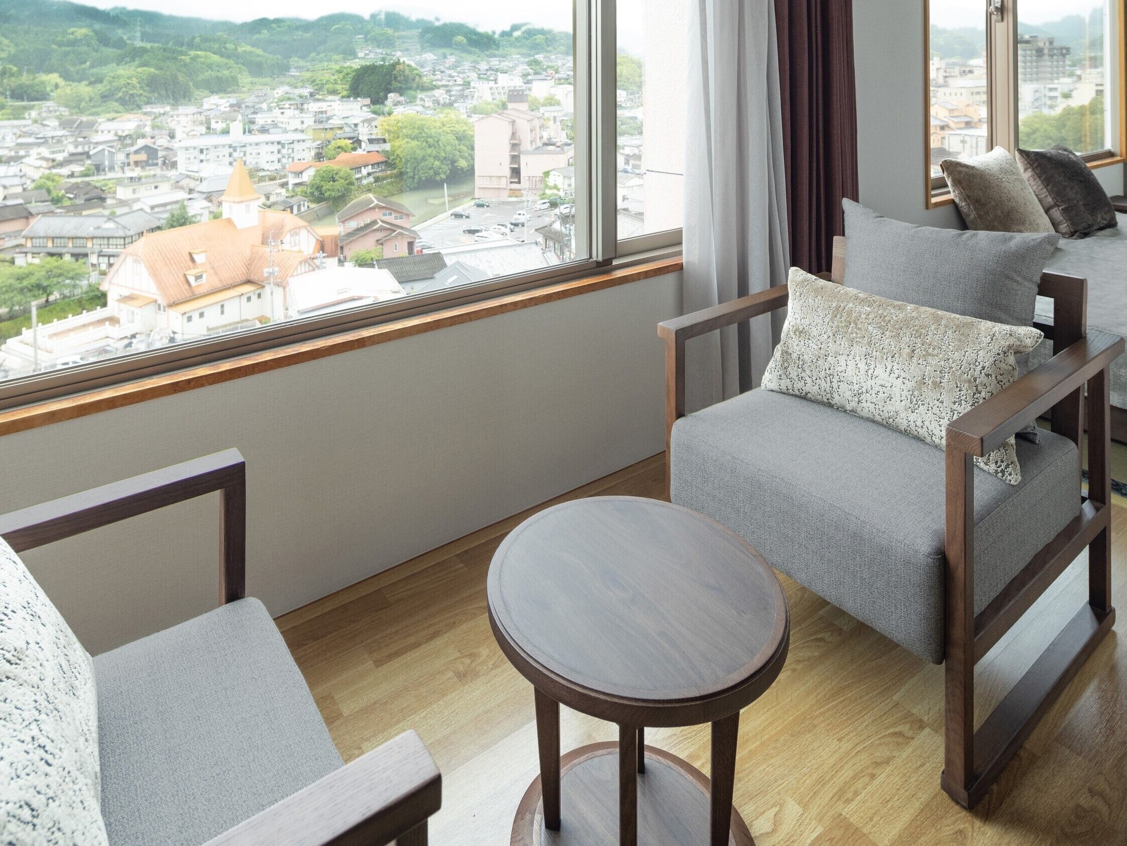 Een moderne kamer met uitzicht op een stadsbeeld door grote ramen. Er staat een bank met houten frame en grijze kussens, en een klein rond tafeltje in het midden.