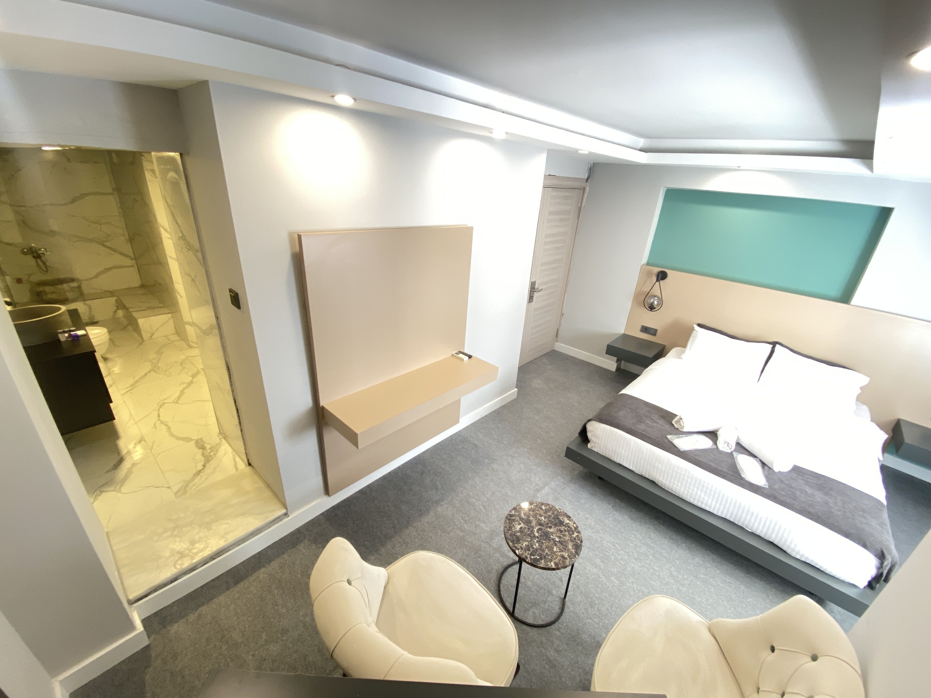 A modern hotel room with a bed, a seating area with chairs, a small table, a vanity with a mirror, and a bathroom with a glass shower enclosure.