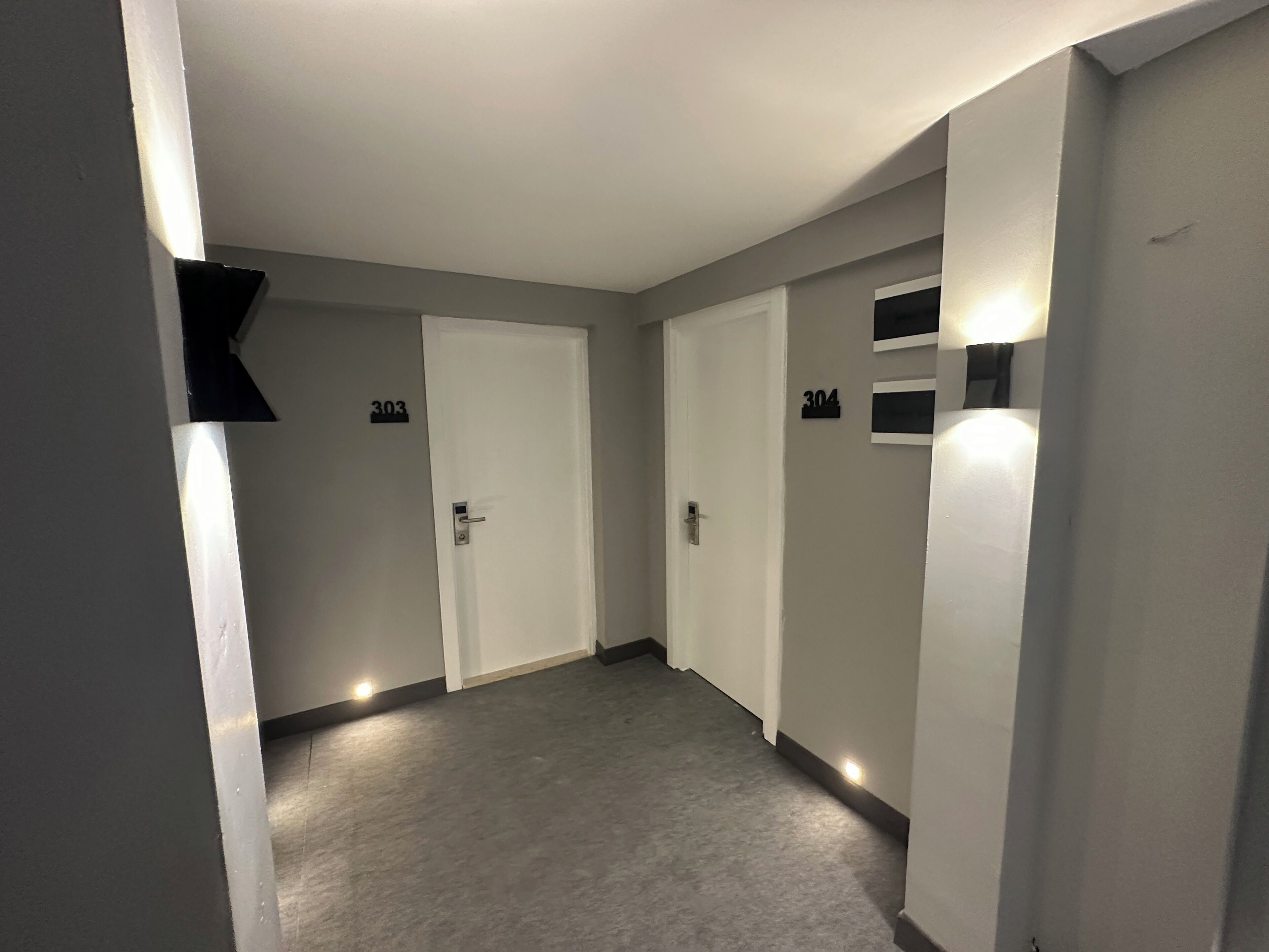A hallway with two doors, each marked with the number 303. The hallway has a grey carpet and is illuminated by wall-mounted lights.