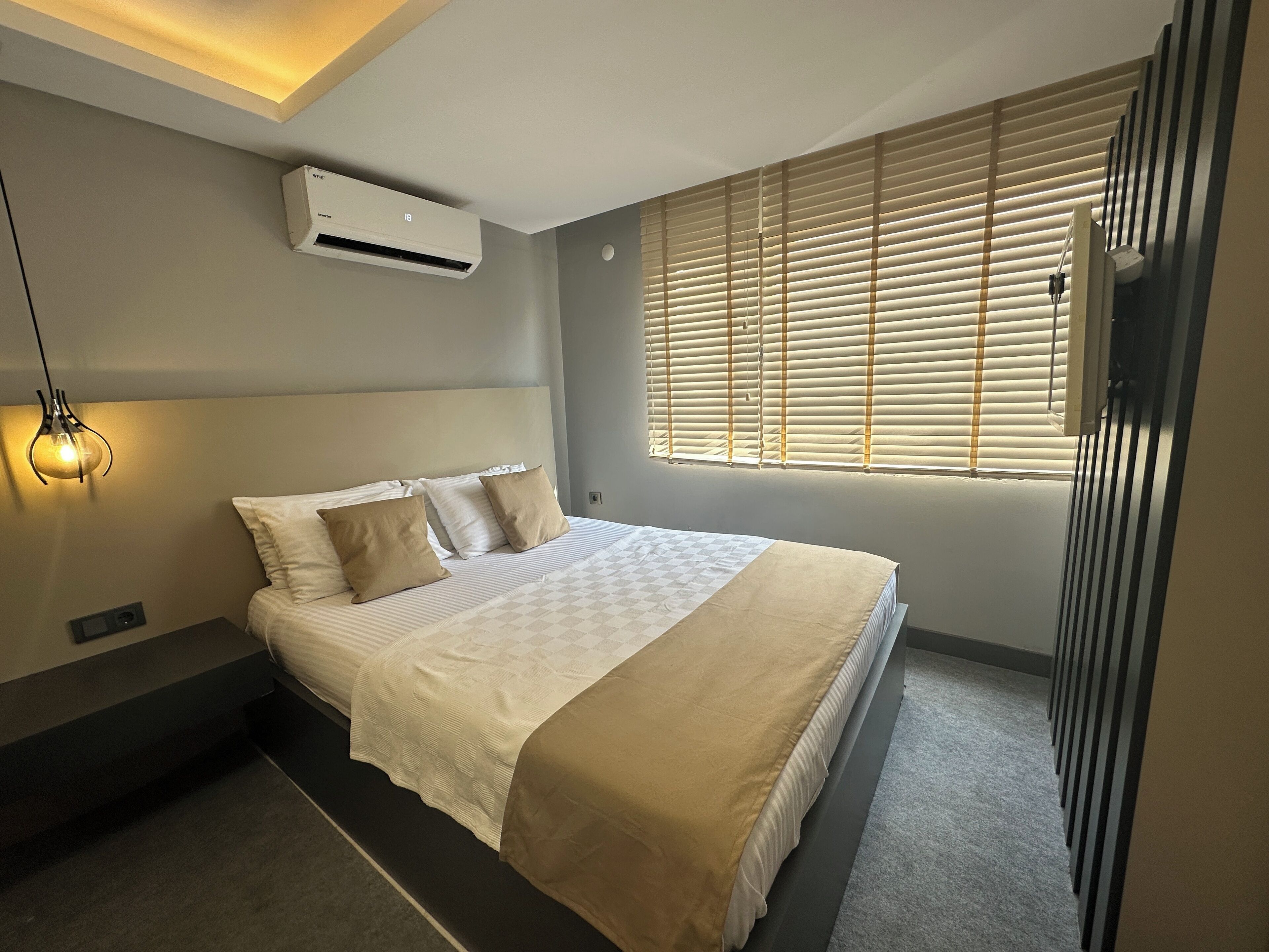 Un dormitorio moderno con una cama grande, un equipo de aire acondicionado, una lámpara de noche y una ventana con persianas.