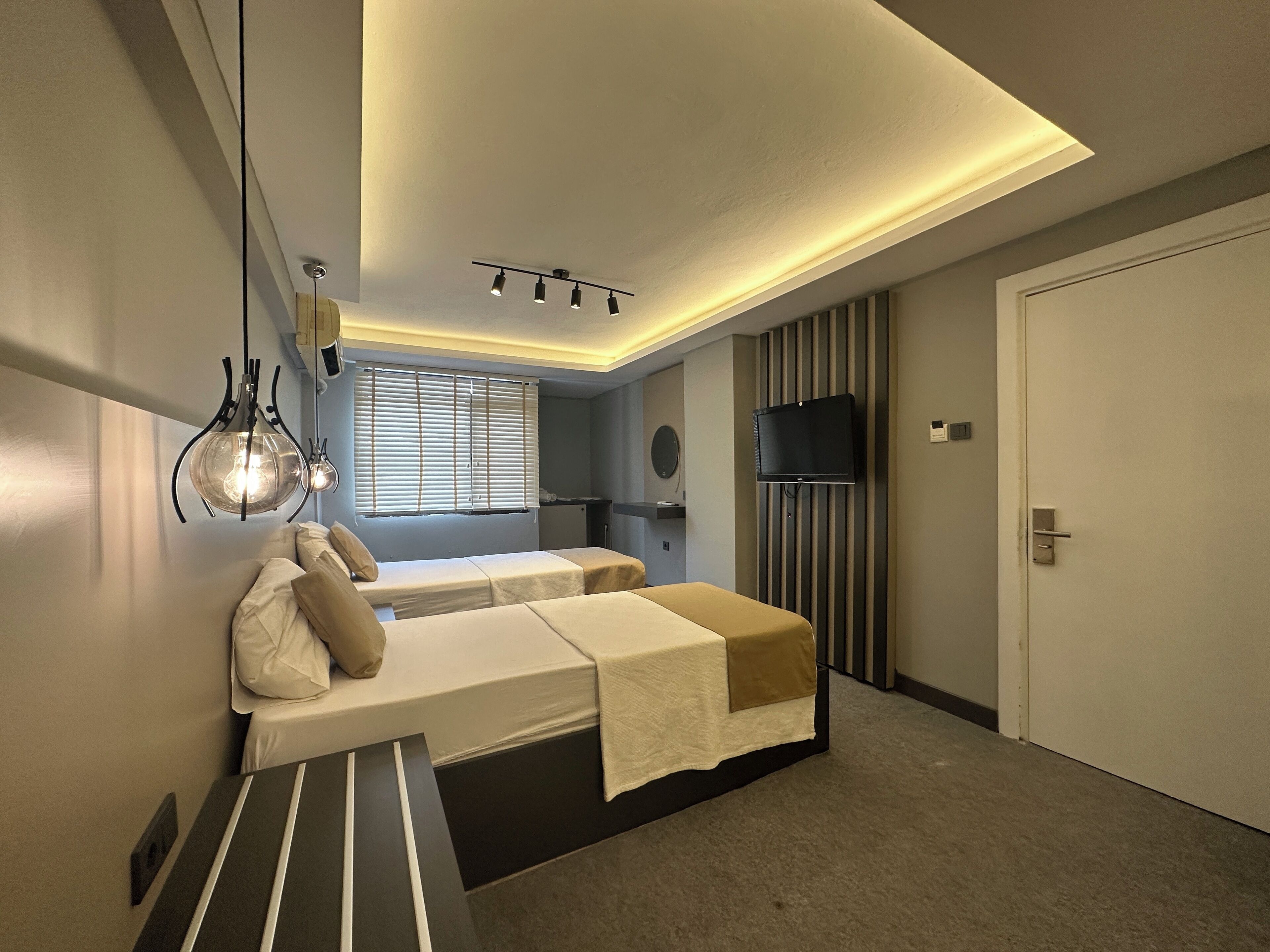 Et moderne hotelværelse med to enkeltsenge, et fladskærms-tv monteret på væggen, en loftlampe og et vindue med persienner.