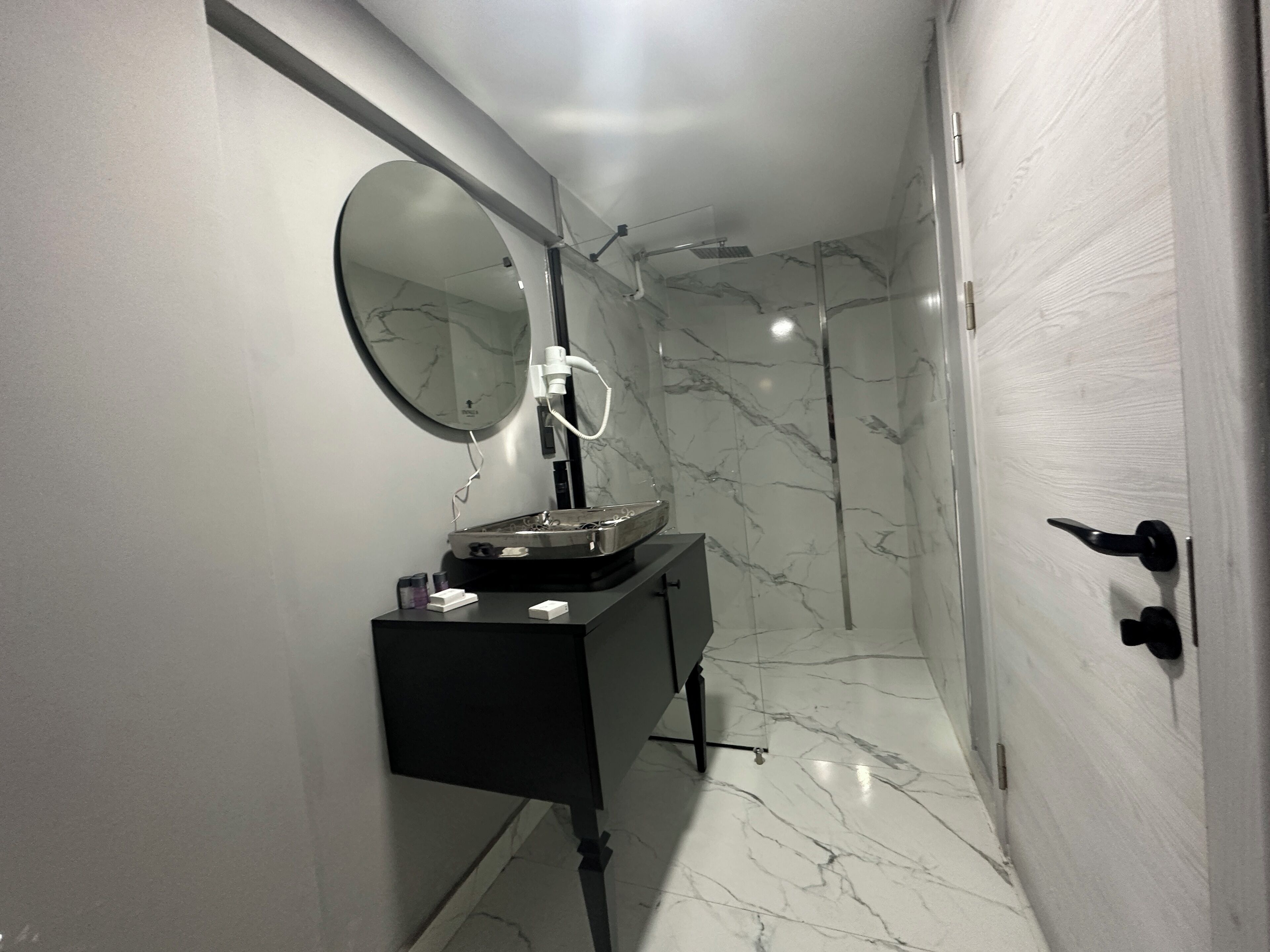 A modern bathroom with a marble floor, a round mirror, a black vanity, and a shower area.