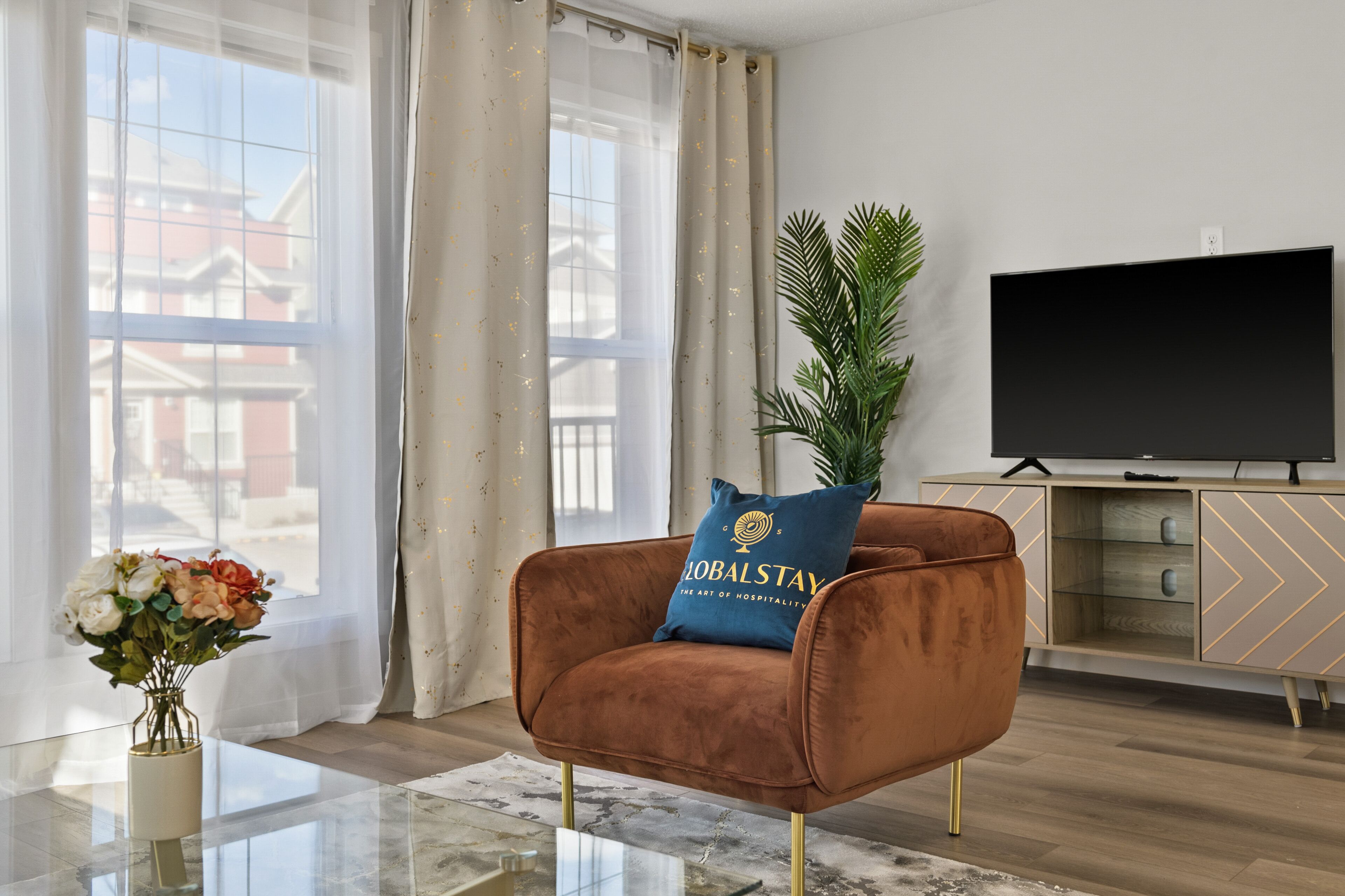 A modern living room with a brown velvet armchair, a flat-screen TV, a glass coffee table, and a potted plant.