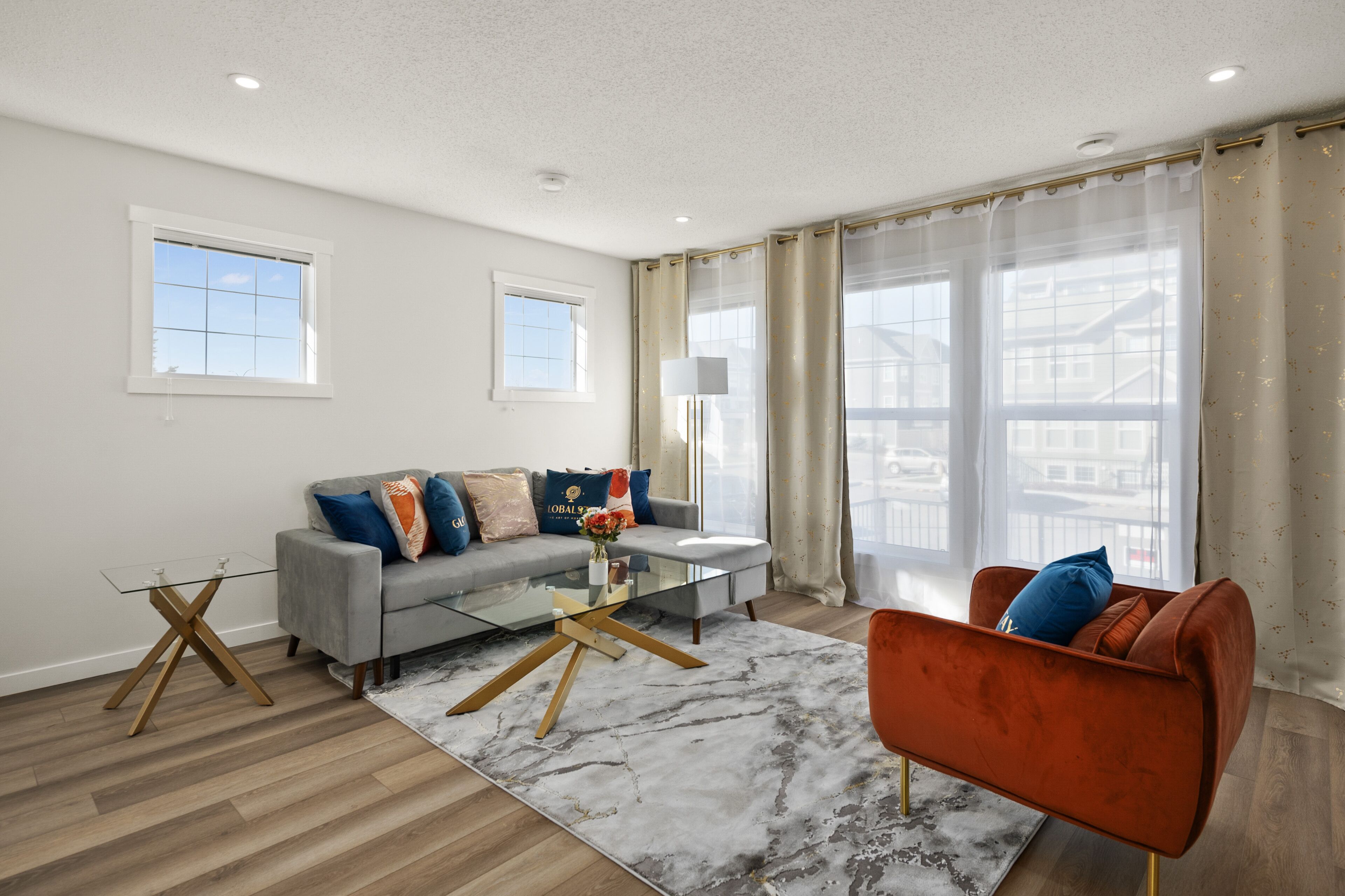 A modern living room with a grey sofa, a glass coffee table, and a red armchair. There are two windows with curtains, a lamp, and a view of buildings outside.