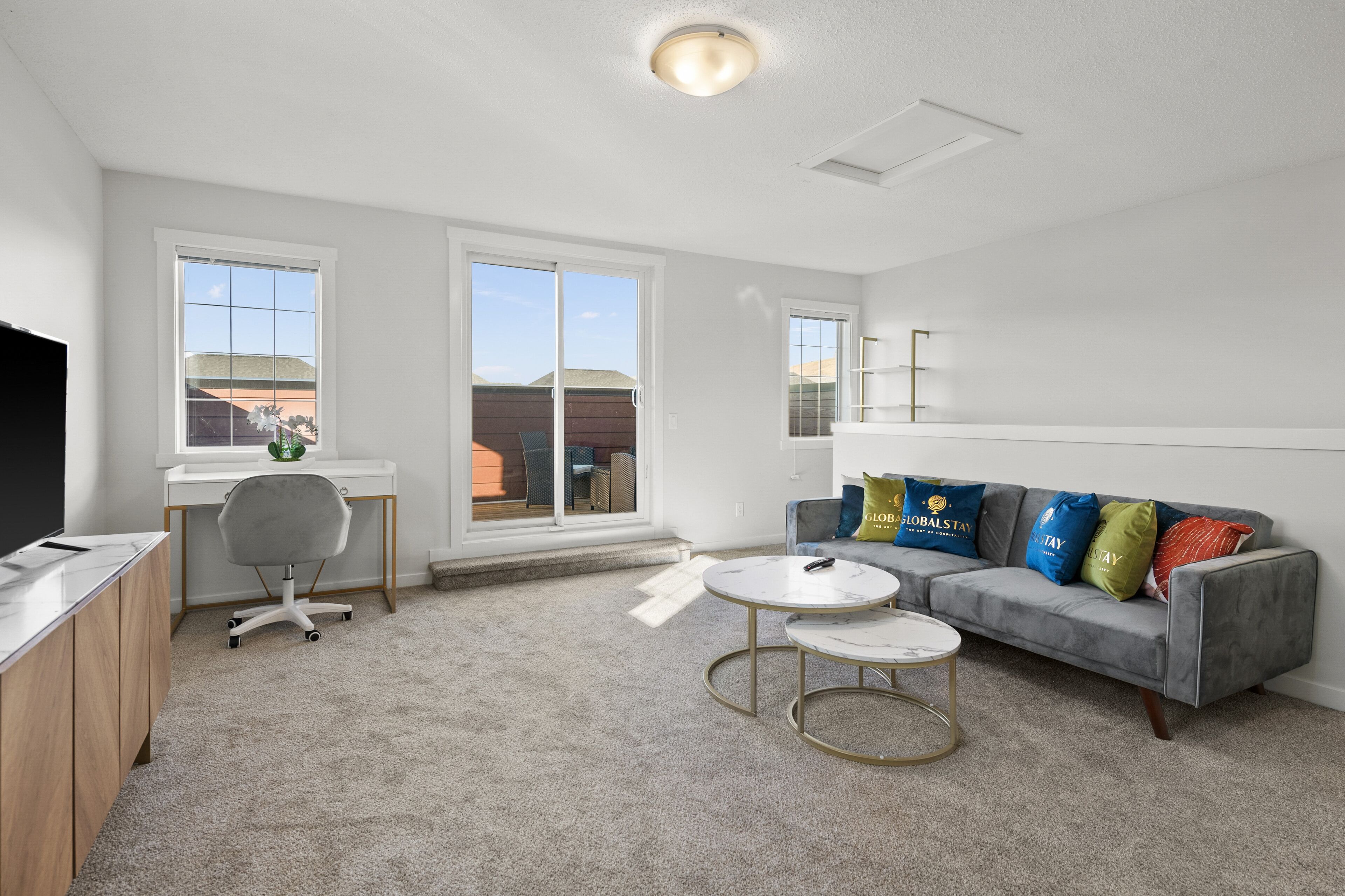 A modern living room with a grey sofa, a white desk, and a grey office chair. There are two windows providing natural light and a view of the outside.