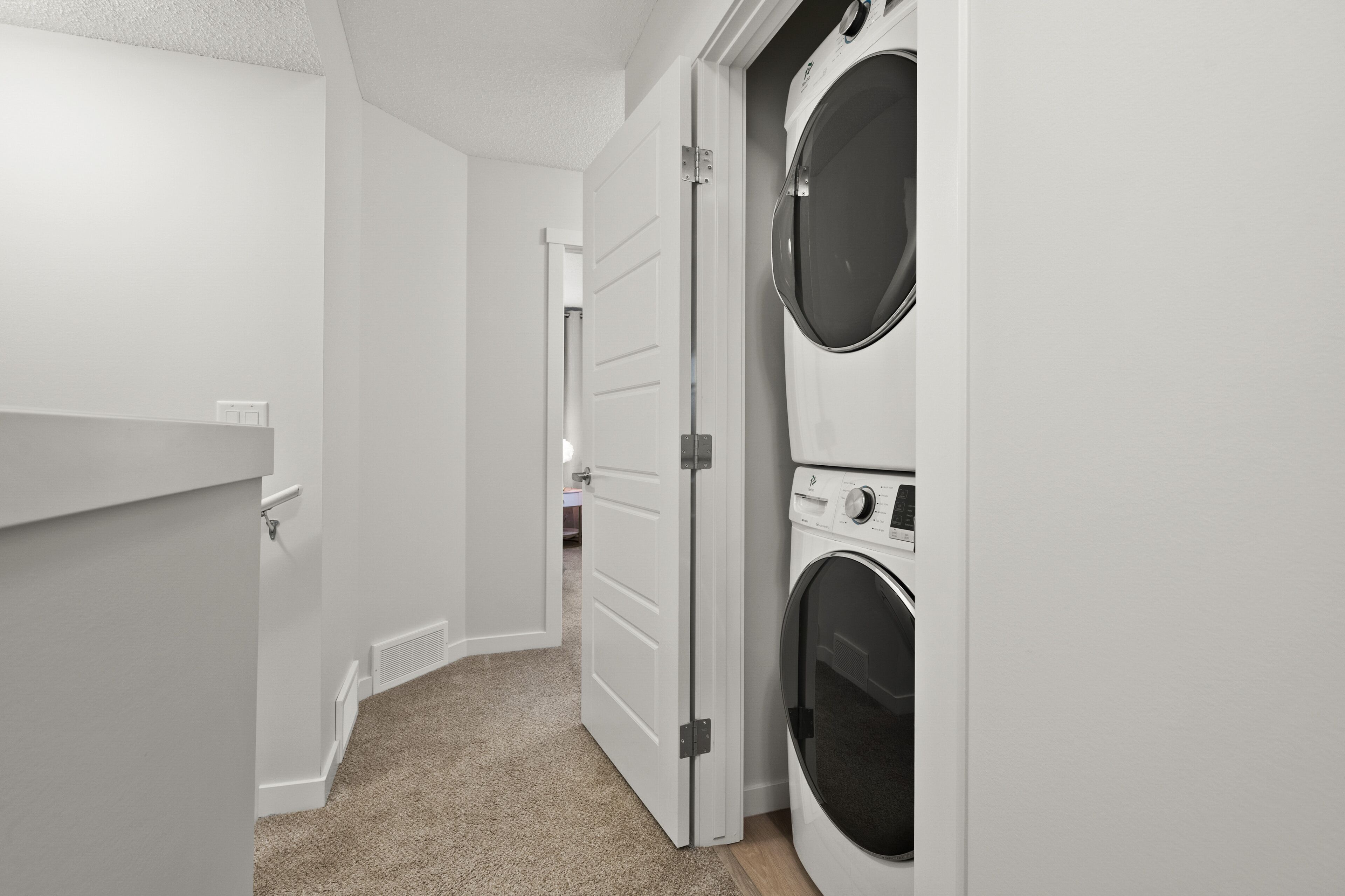 A laundry room with two stacked front-loading washing machines, a white door, and a beige carpet.