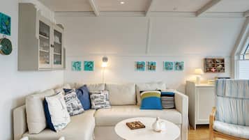 Una sala de estar con un sofá, una mesa de centro, una mesita auxiliar y una silla. Hay estantes montados en la pared con objetos decorativos y una lámpara.