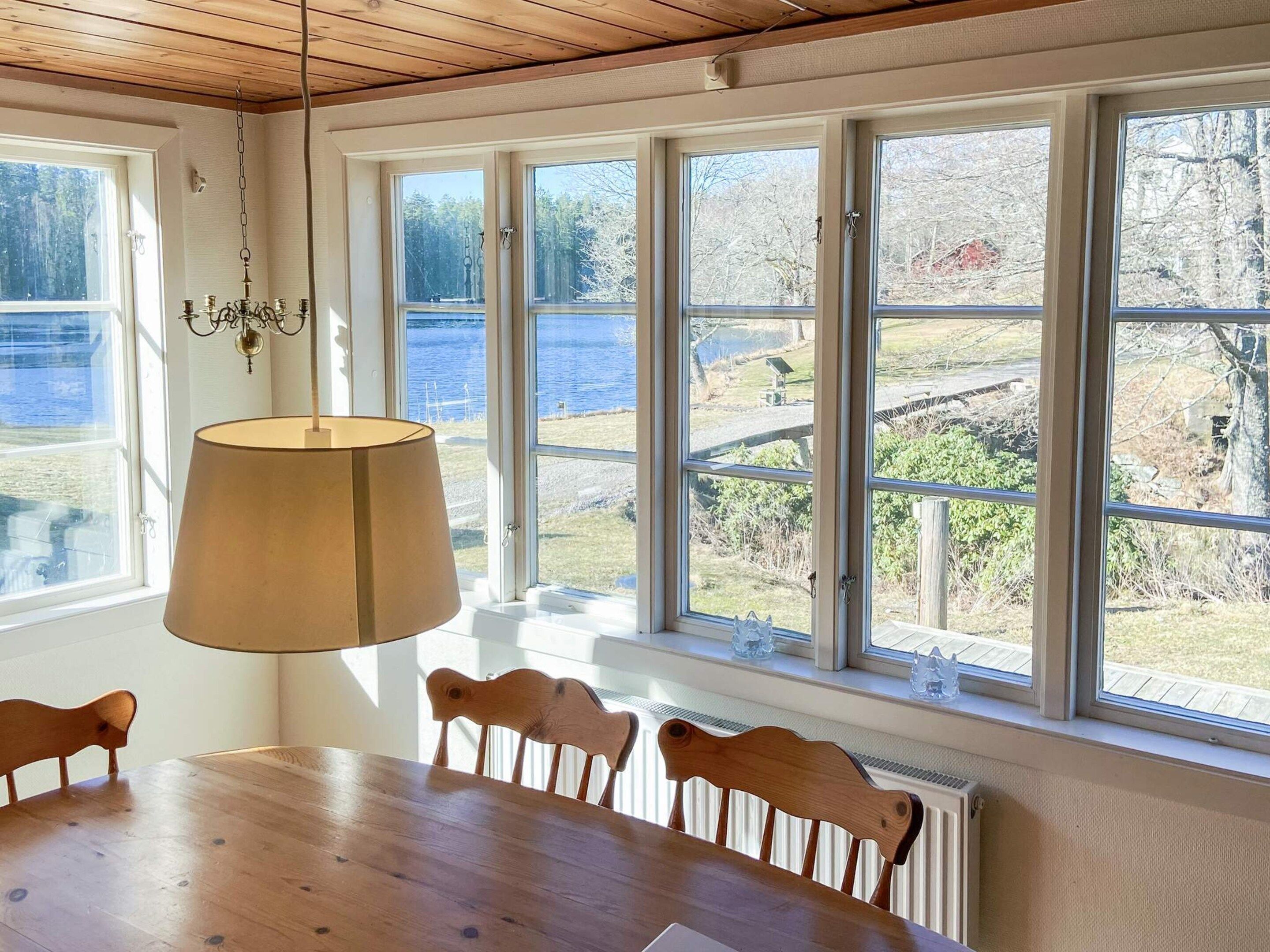 A dining area with a wooden table and chairs, large windows with a view of a lake and trees, and a hanging lamp.