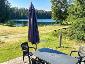 A deck with a table and chairs overlooking a lake, surrounded by trees and a grassy area. - 5 Star Holiday Home in Asbro (Asbro)