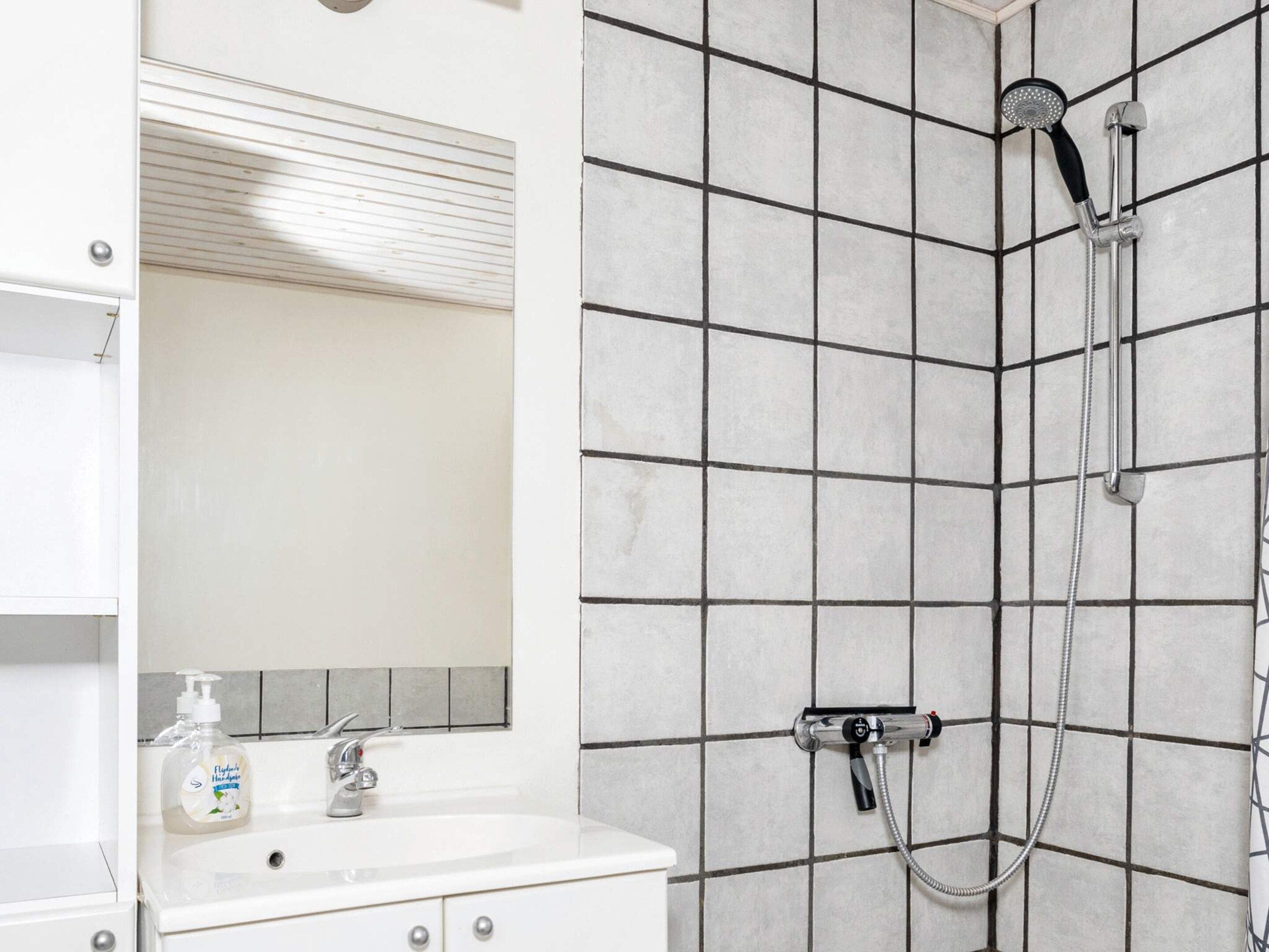 A bathroom with a tiled shower wall, a sink with a soap dispenser, and a mirror above the sink.