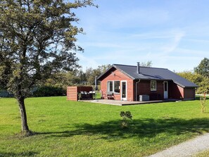 A red wooden house with a covered patio, a small garden, and a tree in the foreground.