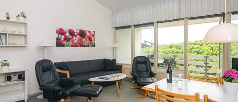Een moderne woonkamer met een bank, twee leren fauteuils, een eettafel met stoelen, een salontafel en een groot raam met uitzicht op groen.