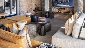 Ein großzügiges Wohnzimmer mit hohen Decken, freiliegenden Backsteinwänden, einem großen Fernseher und bequemen Sitzgelegenheiten.