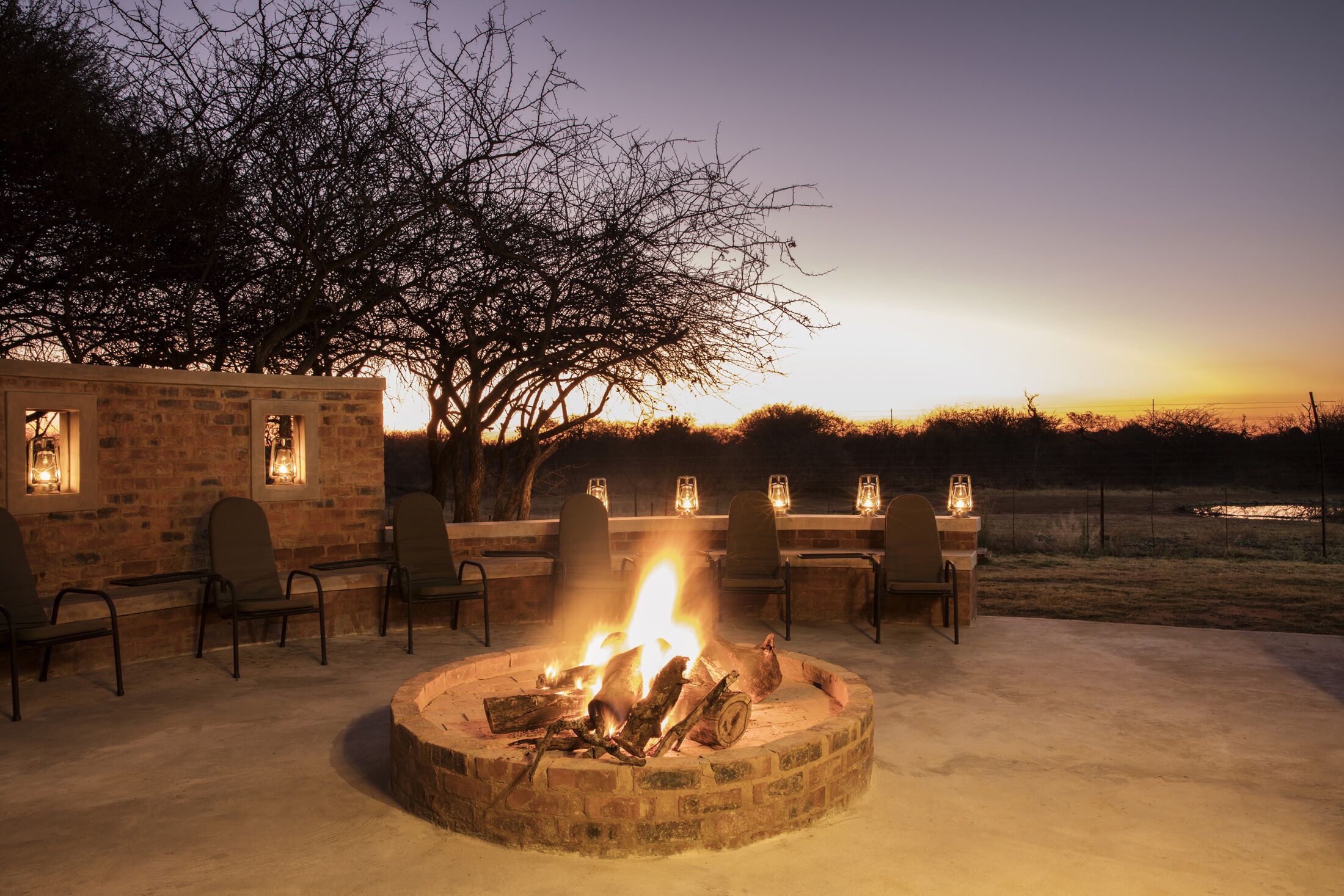 A circular stone fire pit with logs, surrounded by chairs and tables, set against a backdrop of a tree and a sunset.