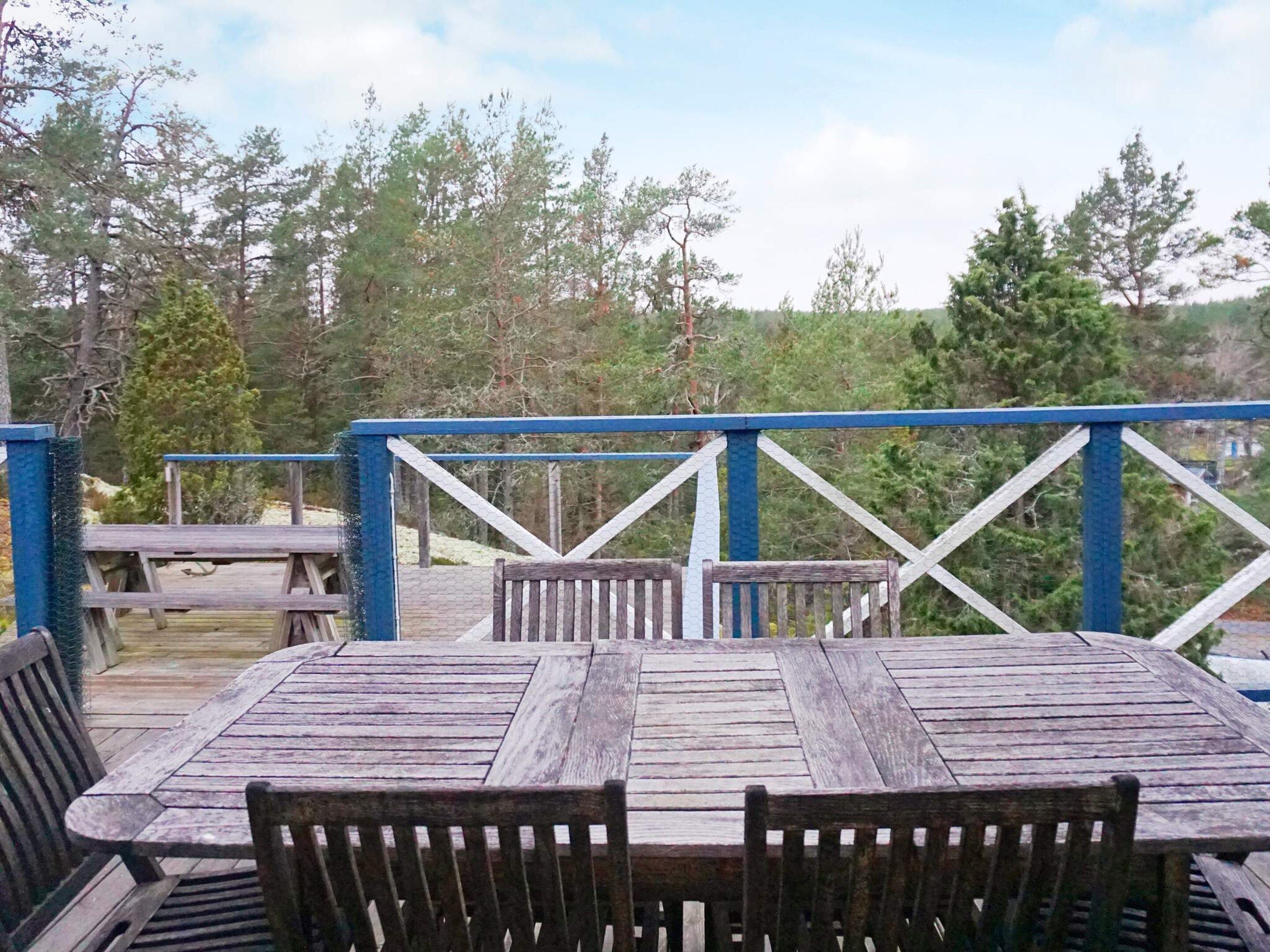 Een houten terras met een tafel en stoelen, omringd door een blauwe balustrade, met uitzicht op een bebost gebied.