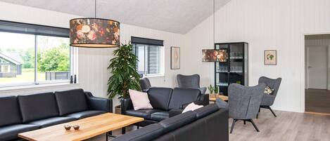 A modern living room with a sectional sofa, a coffee table, and a dining table. There is a large window providing natural light and a view of the outside.