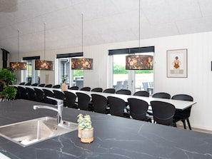 Une salle de conférence moderne, dotée d’une longue table et de chaises, d’un îlot en marbre avec un évier, et de grandes fenêtres équipées de stores.