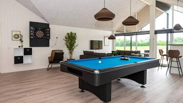 Een moderne biljarttafel binnen met een blauwe speelbaan, omringd door een houten vloer, een aan de muur bevestigde televisie en een groot raam met uitzicht op groen.