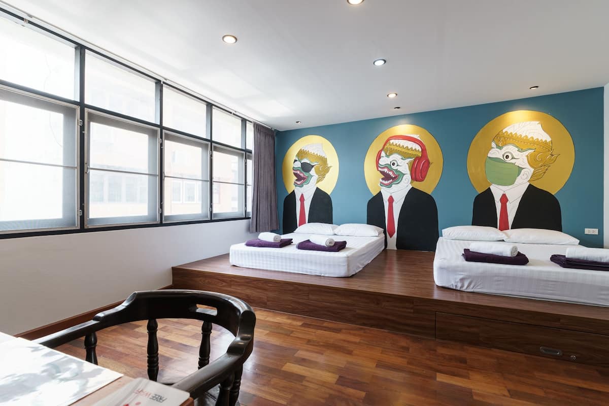 A hotel room with two beds, wooden flooring, a desk with a chair, and a large window.