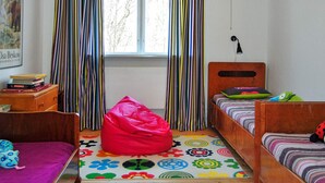 Une pièce avec deux lits en bois, un fauteuil en forme de poire, un tapis coloré, une fenêtre avec des rideaux et une lampe fixée au mur.