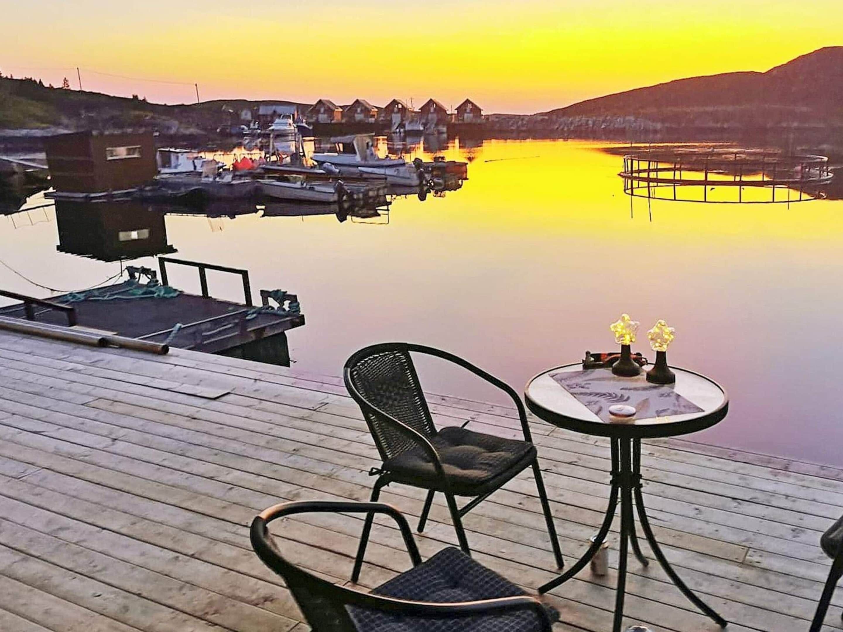 Una terraza de madera con una mesa y sillas, con vista a una masa de agua tranquila donde se ven barcos y casas al fondo.