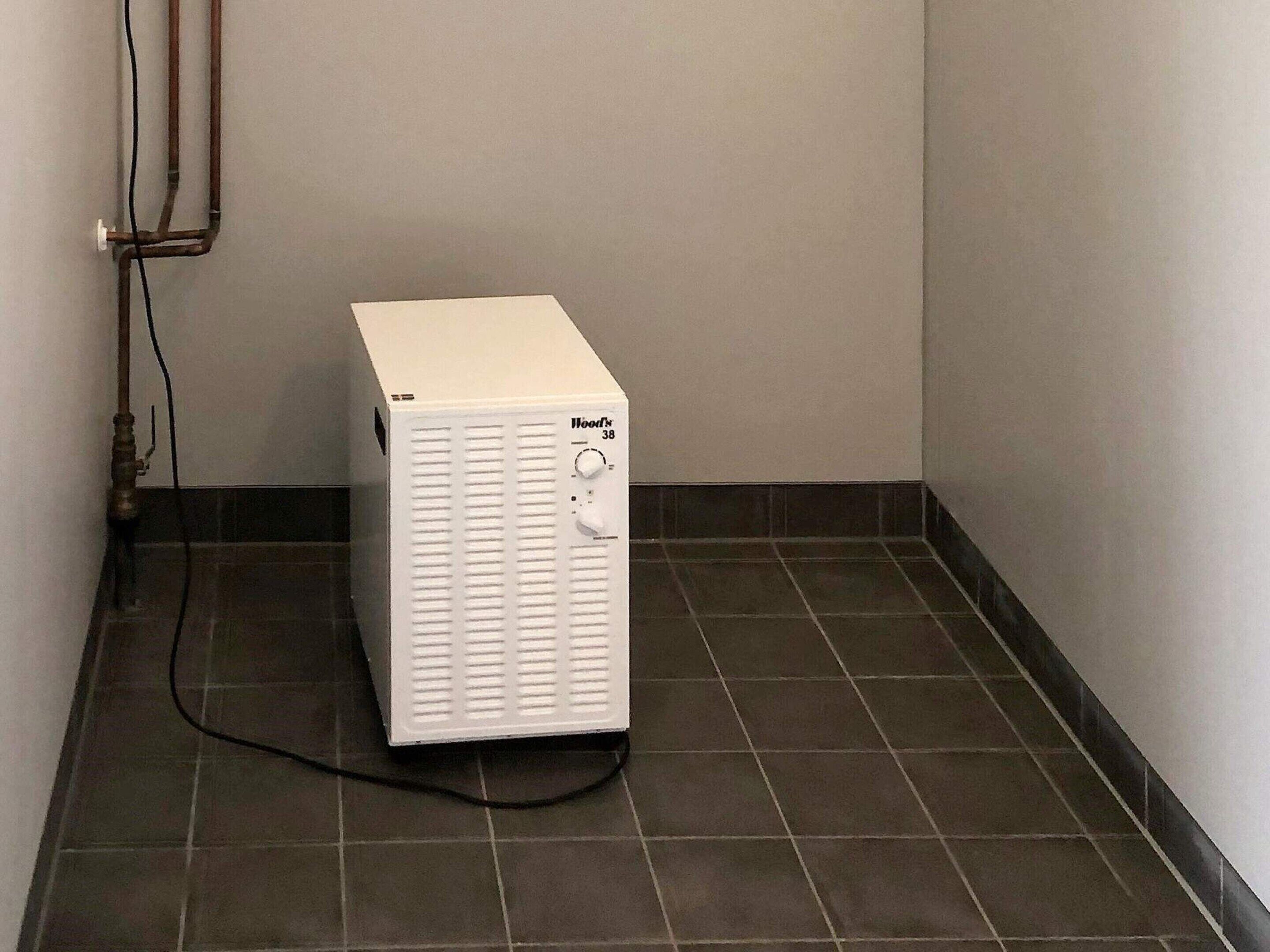 A white portable heater labeled "Hoot's 38" in a corner with tiled flooring and a wall-mounted pipe.