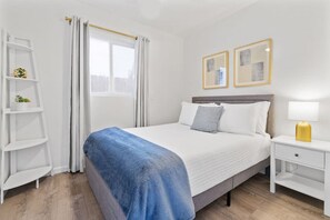 A neatly arranged bedroom with a bed, bedside tables, a shelf with a plant, and a window with curtains. - Coastal 2BR Escape with High-Speed WiFi (San Diego)