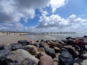A beach with large rocks in the foreground, a pier extending into the water, and people walking along the shore. - 4 Person Holiday Home in Blavand (Blavand)