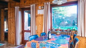A wooden interior with a dining table covered in a colorful tablecloth, chairs, a bottle of wine, and a view of greenery outside.