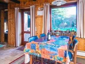 A wooden interior with a dining table covered in a colorful tablecloth, chairs, a bottle of wine, and a view of greenery outside.