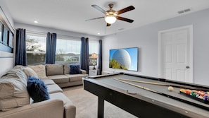 Uma sala de estar com mesa de bilhar, sofá, ventoinha no teto e uma grande janela com cortinas.
