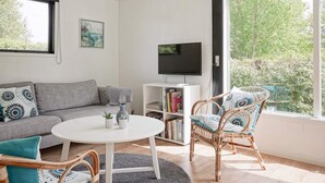 A modern living room with a grey sofa, a round white table, wicker chairs, a bookshelf, and a TV. - 6 Person Holiday Home in Allinge (Allinge)