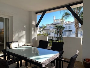 A balcony with a glass table and chairs, overlooking a white building with palm trees.