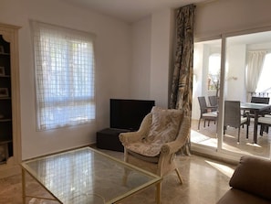 A living room with a glass coffee table, a wicker armchair, a flat-screen TV, and a sliding glass door leading to a dining area.